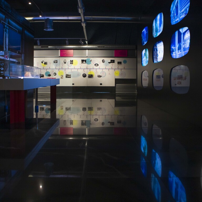 Un suggestivo itinerario multimediale in una sala buia con schermi accesi, teche e una linea del tempo sulla parete di fondo.