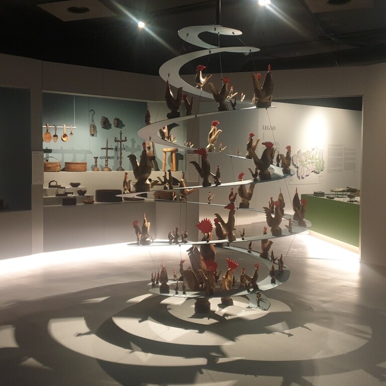 Una grande spirale con galletti di legno si erge al centro di una sala museale, davanti a una parete con taglieri e utensili d'epoca.