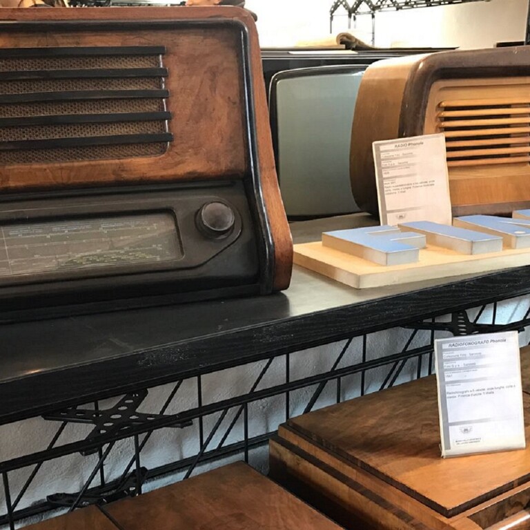Due radio d'epoca in legno con griglie frontali sono esposte su uno scaffale espositivo insieme a schede informative.