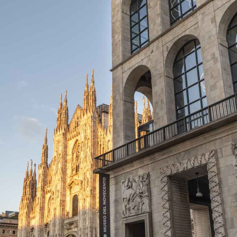 Palazzo monumentale in pietra con ampie arcate in primo piano, affacciato su una cattedrale gotica illuminata dal sole.