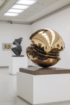 Luminoso spazio espositivo con opere d'arte: in primo piano una scultura sferica in bronzo lucido con una superficie spaccata e complessi ingranaggi interni.