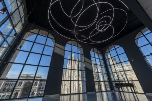 Ampia sala con installazione luminosa a spirale sul soffitto e vetrate ad arco affacciate sul Duomo gotico.