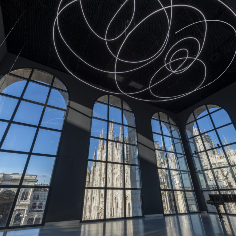 Ampia sala con installazione luminosa a spirale sul soffitto e vetrate ad arco affacciate sul Duomo gotico.