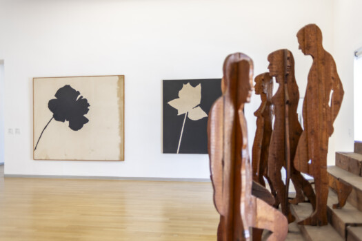 Spazio espositivo essenziale con sculture lignee sagomate a figura umana lungo una scala e quadri minimalisti a parete.
