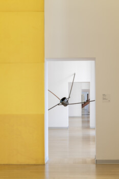 Spazio museale moderno con parete gialla e porte in sequenza prospettica che inquadrano una scultura metallica sospesa.