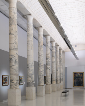 Ampia galleria espositiva scandita da un imponente colonnato in marmo venato, soffitto voltato luminoso e dipinti alle pareti.