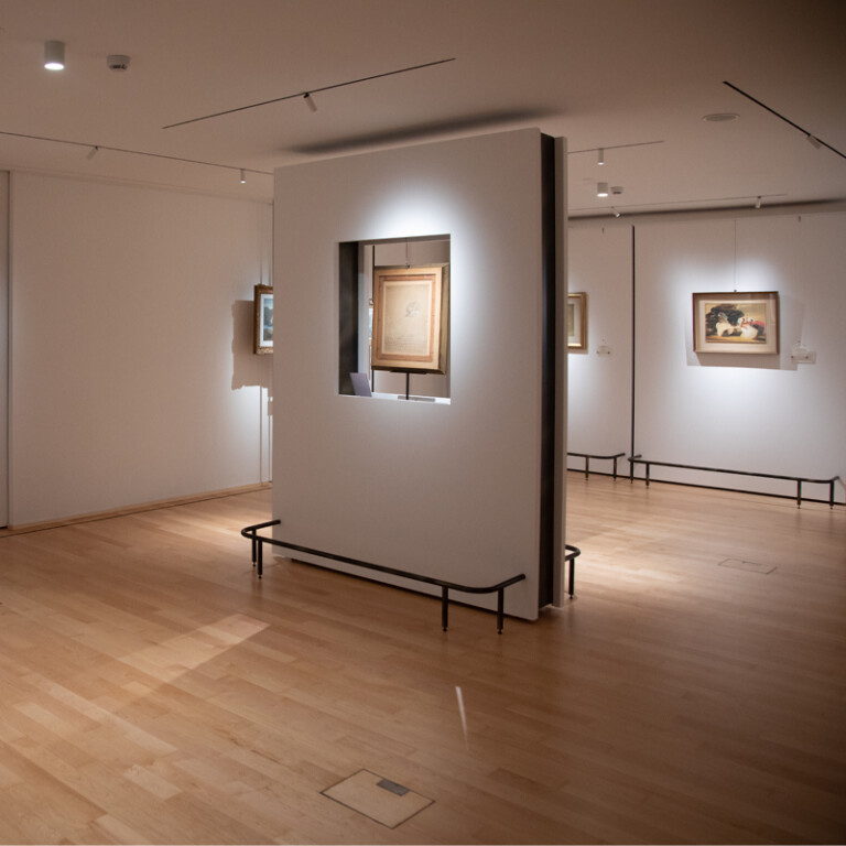 Elegante sala museale con parquet e pareti chiare, dove quadri e stampe sono esposti su pareti e su un pannello divisorio centrale.