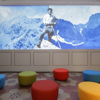 Sala espositiva accogliente con vivaci sedute colorate, dominata da un grande pannello azzurro con la foto di Pier Giorgio Frassati in cima a una montagna.