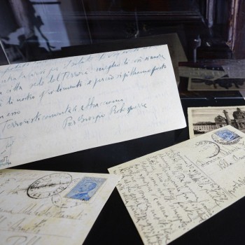 Dettaglio ravvicinato di lettere e cartoline d'epoca fittamente scritte a mano, esposte all'interno di una teca museale.