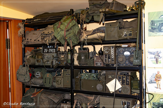 Scaffali metallici ospitano una ricca collezione di radio ricetrasmittenti militari storiche e zaini d'equipaggiamento da campo.