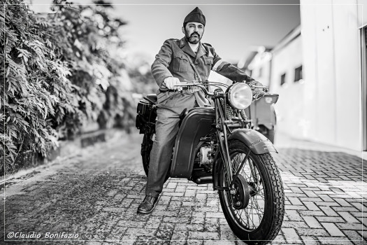 Scatto contemporaneo in bianco e nero di un uomo in uniforme storica su una moto d'altri tempi, lungo un vialetto pavimentato e alberato.