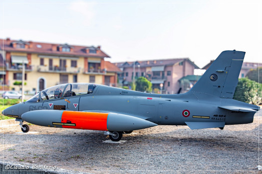 Un aereo militare grigio con serbatoio laterale arancione, esposto all'aperto davanti a edifici residenziali di sfondo.