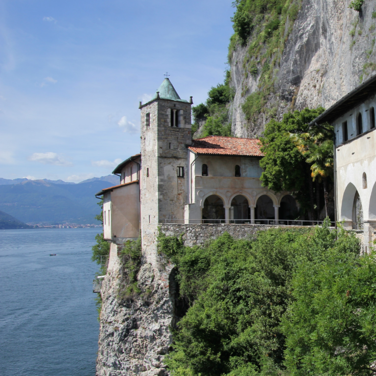 Antico monastero in pietra arroccato su una scogliera a picco sul lago con un campanile cuspidato e un elegante porticato ad archi immerso nel verde.